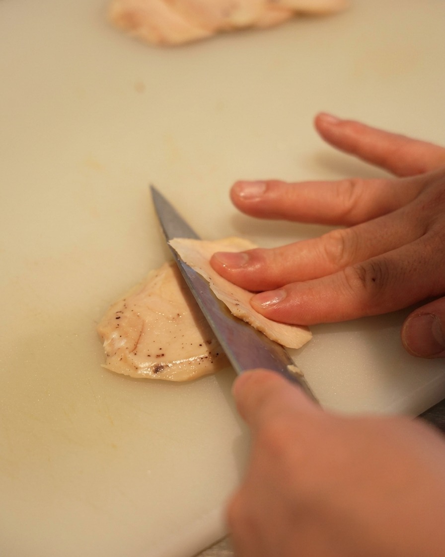 Primo piano di una mano che usa un coltello affilato per affettare un petto di pollo chiaro in pezzi sottili su un tagliere bianco.