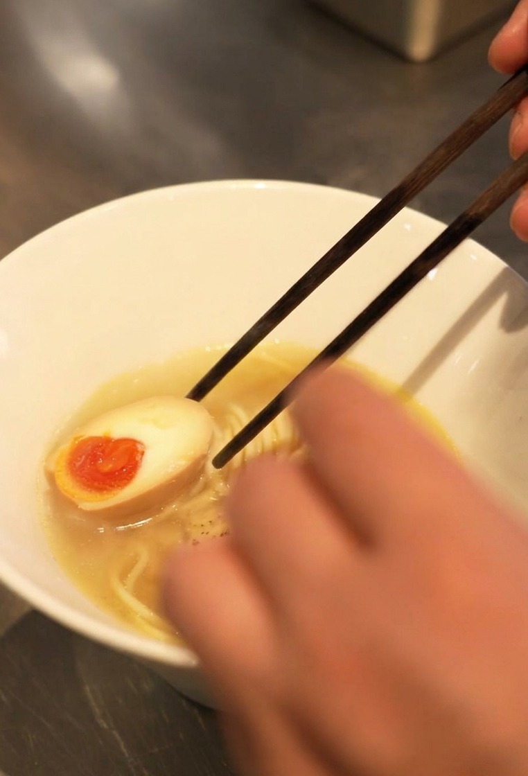 Dettaglio di bacchette che inseriscono mezzo uovo sodo con un tuorlo arancione brillante e cremoso in una ciotola di ramen.