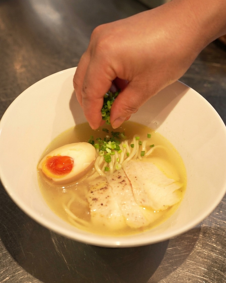 Una mano cosparge cipollotti freschi tritati su una ciotola completa di ramen shio al pollo con uovo e fette di pollo.