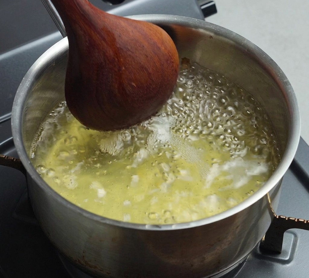 Una pentola di acciaio inossidabile piena di brodo chiaro che bolle, mescolato con un cucchiaio di legno scuro.