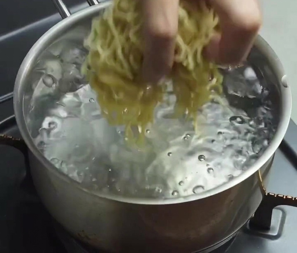 Una mano che cala un mazzetto di noodles ramen freschi, sottili e gialli, in una pentola dacqua limpida che bolle rapidamente.