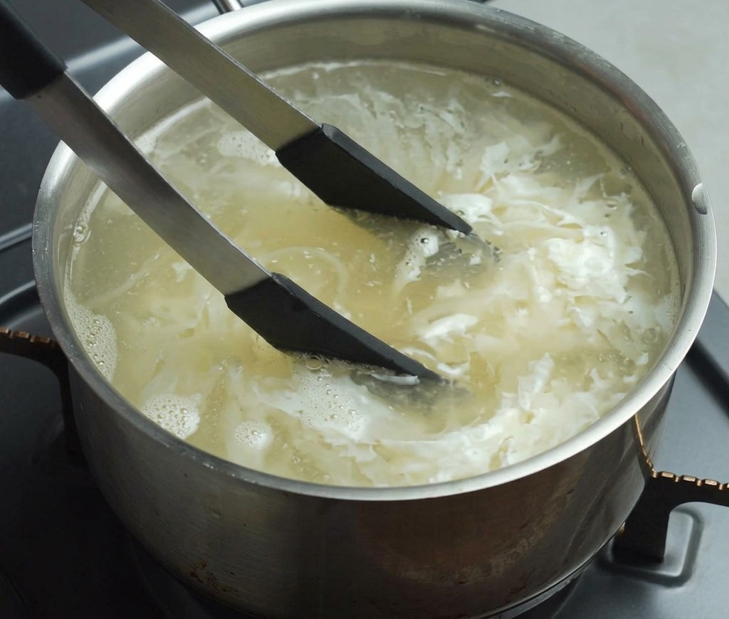 Pinze con la punta nera che mescolano noodles gialli in una pentola di acqua bollente sul fornello.