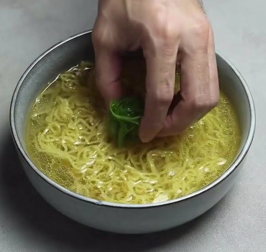 Una mano che posiziona un ciuffo di cipollotto tagliato a listarelle sottili sui noodles ramen in una ciotola di brodo giallo limpido.