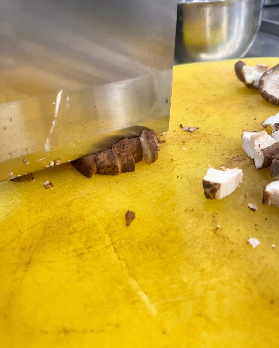 Un coltello da cucina affilato taglia i cappelli dei funghi shiitake a cubetti su un tagliere giallo.