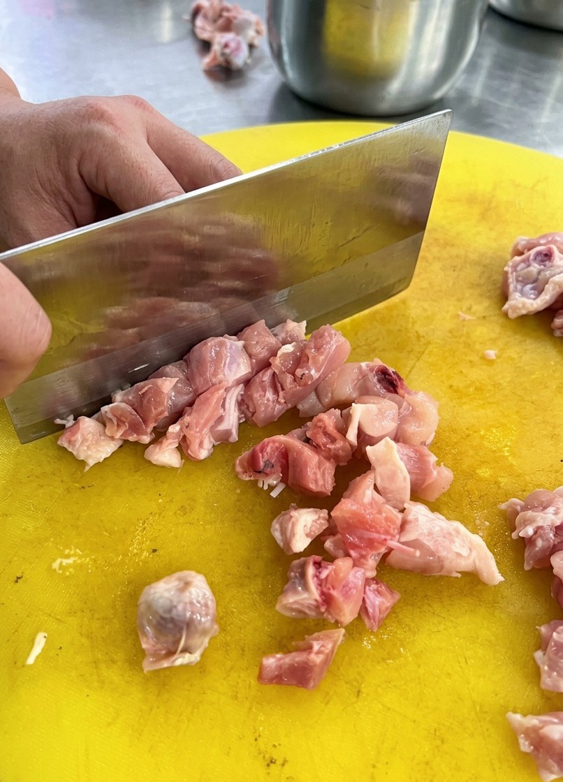 Una mannaia da cucina taglia il petto di pollo crudo a cubetti regolari su un tagliere di plastica giallo.