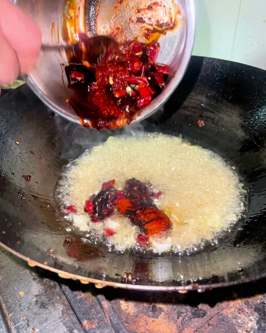 Una densa salsa rossa versata da una ciotola in un wok contenente olio caldo e aromi.