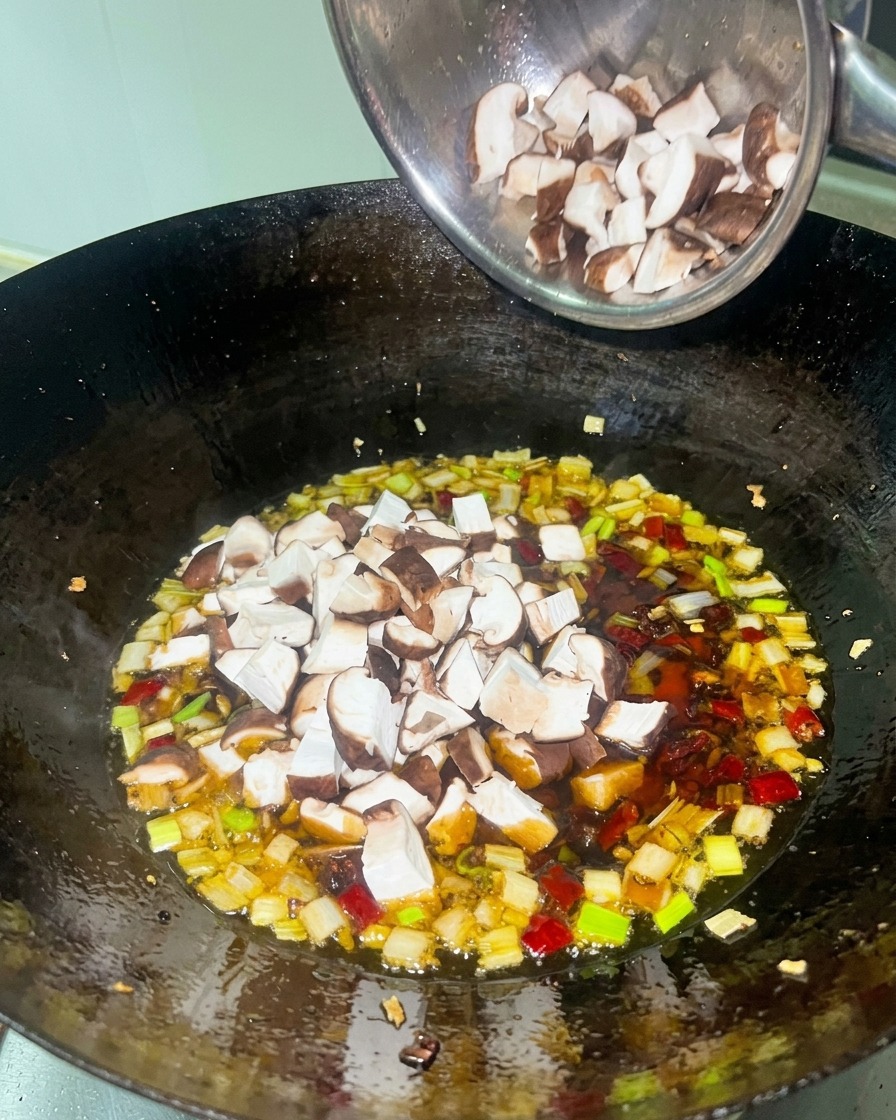 Cubetti di pollo e funghi shiitake aggiunti a una salsa rossa ricca in un wok nero.