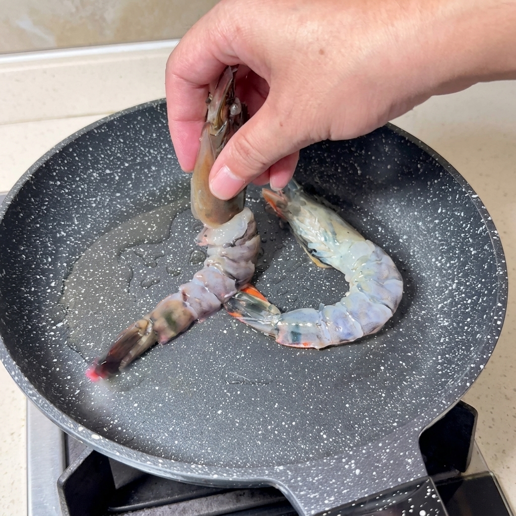 Una mano che adagia grandi gamberi crudi a farfalla in una padella calda grigia maculata con olio scintillante.