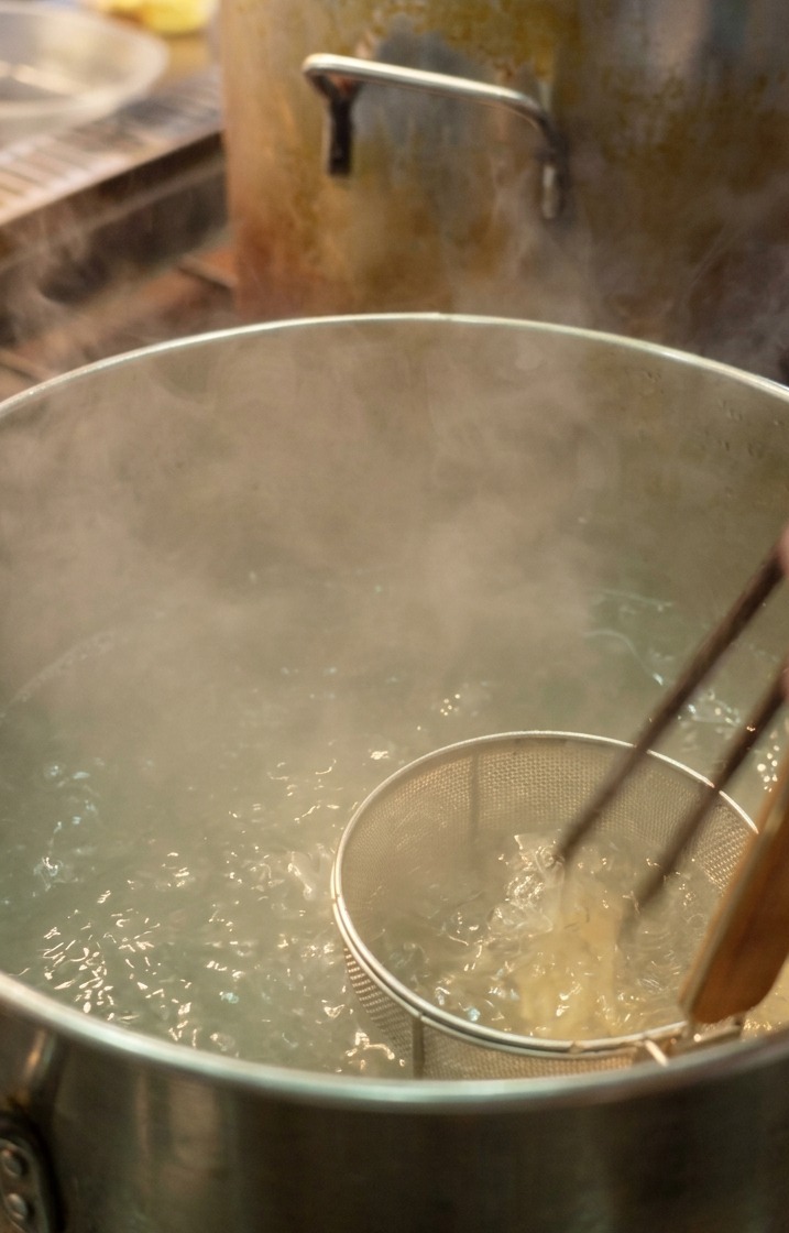 Uno chef che usa un colino a rete per immergere sottili noodles bianchi in una grande pentola di acqua bollente e fumante.