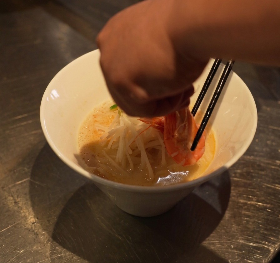 Una mano che usa le bacchette per posizionare un gambero cotto sopra un letto di noodles e germogli di soia.
