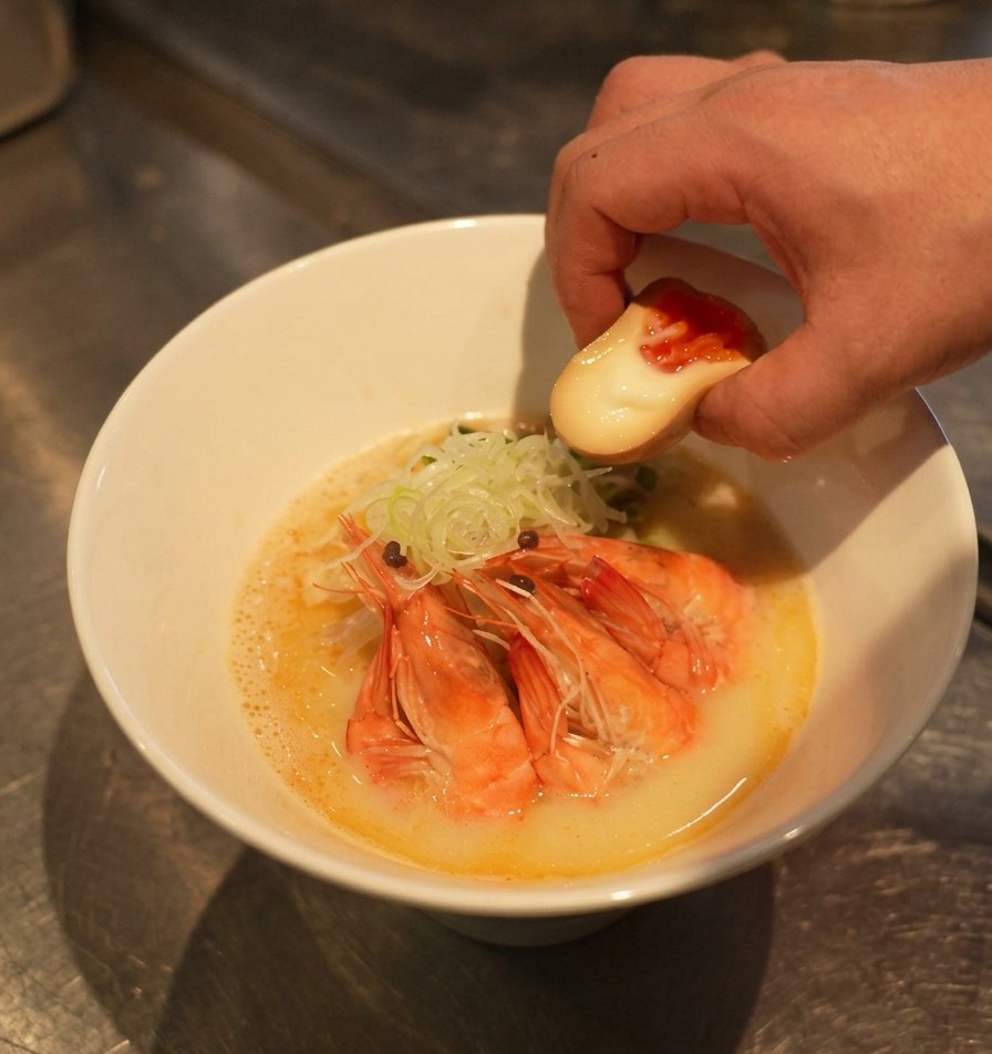 Una mano che posiziona mezzo uovo sodo con il tuorlo arancione lucido in una ciotola di ramen già guarnita.