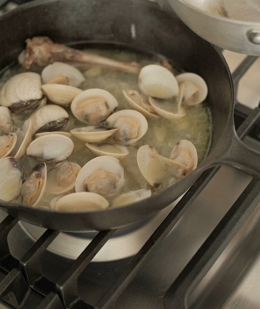 Vongole sollevate da una padella di ghisa contenente un osso danatra e brodo saporito con pinze di metallo.