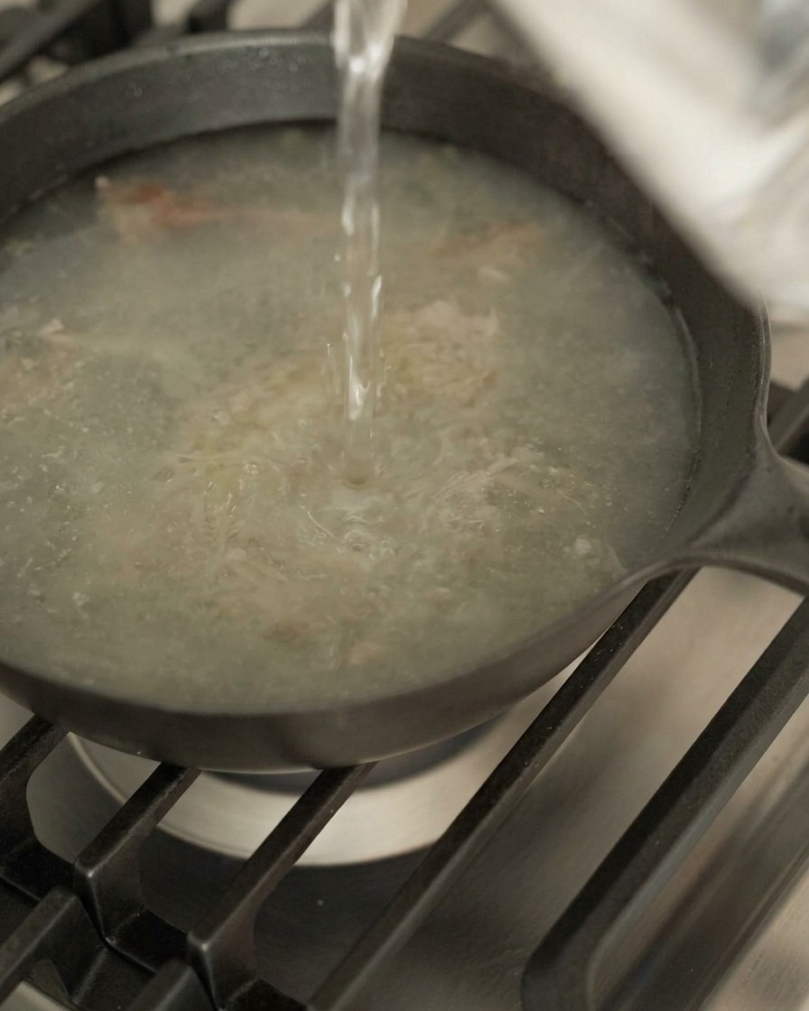 Acqua versata da una certa altezza in una padella scura contenente succhi di carne e aromi.