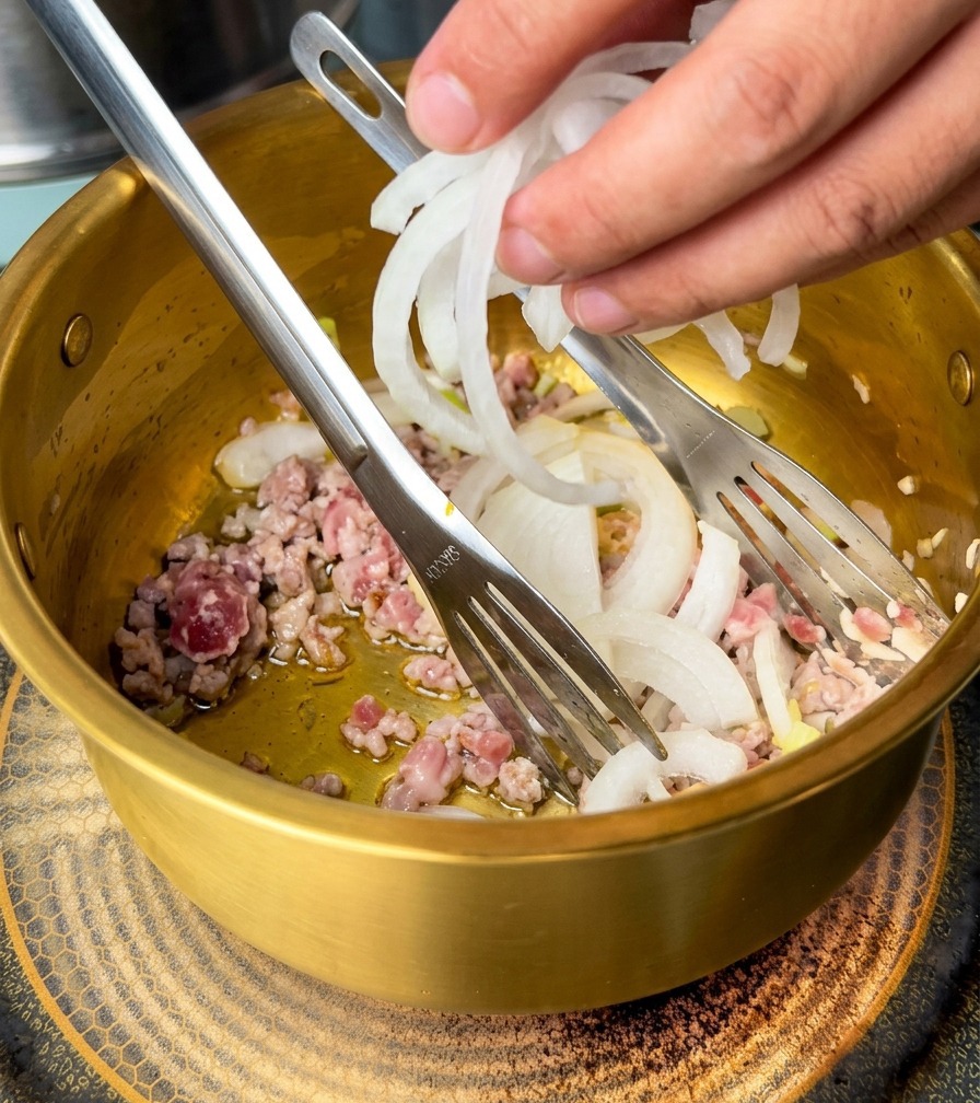 Mano che aggiunge cipolle bianche affettate in una pentola dorata contenente maiale macinato e aromi.