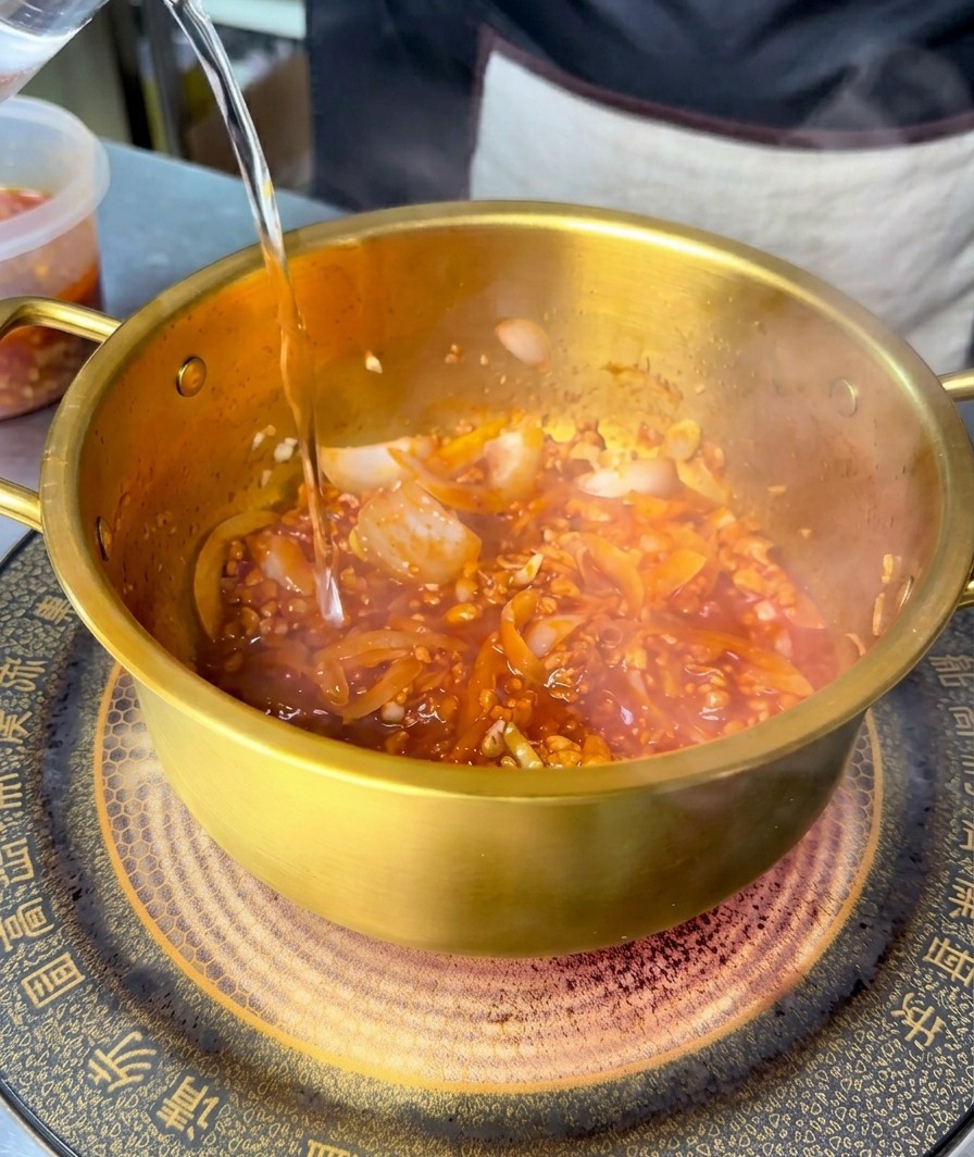 Acqua limpida versata in una pentola dorata contenente una base di carne e verdure rossa e piccante.