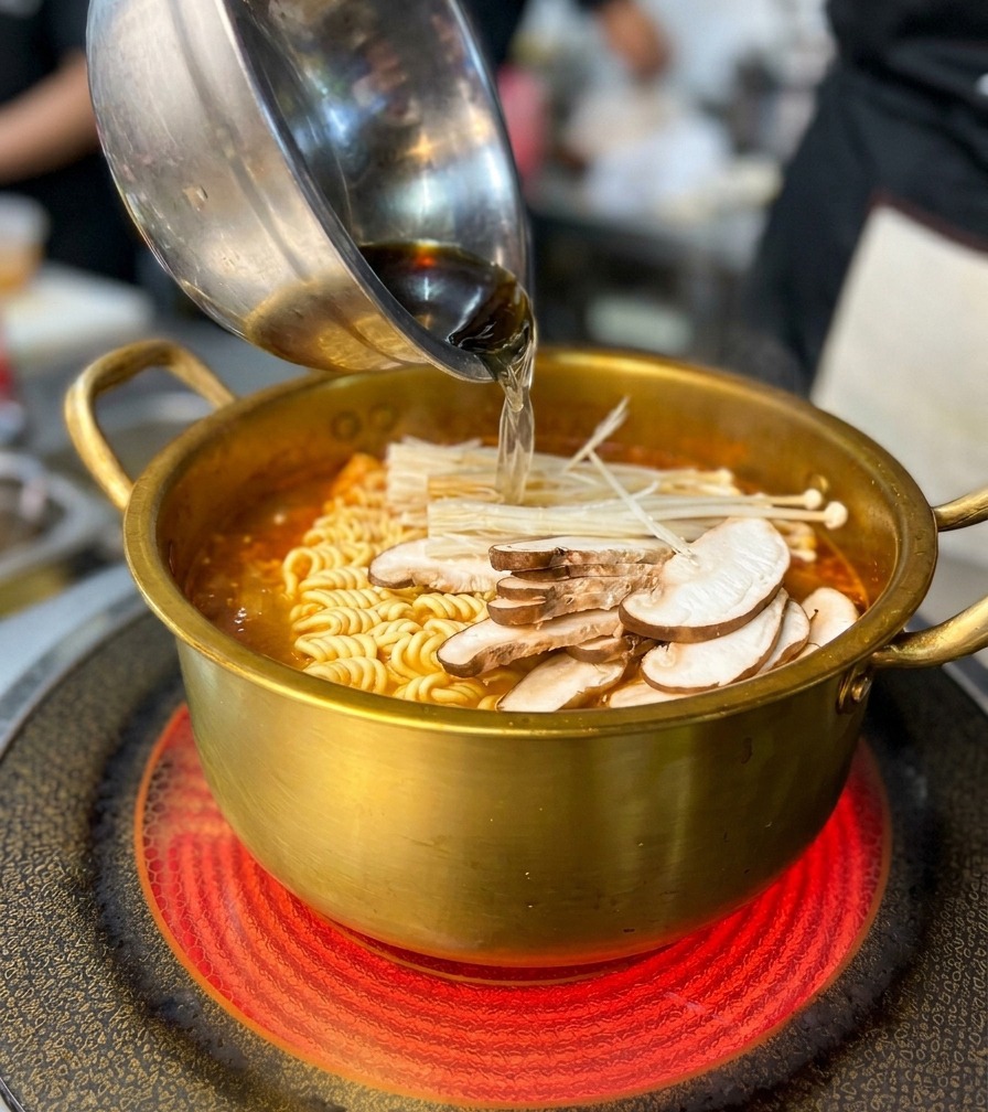Aggiunta di una ciotola di funghi e frutti di mare in una pentola dorata piena di noodle e brodo rosso piccante.