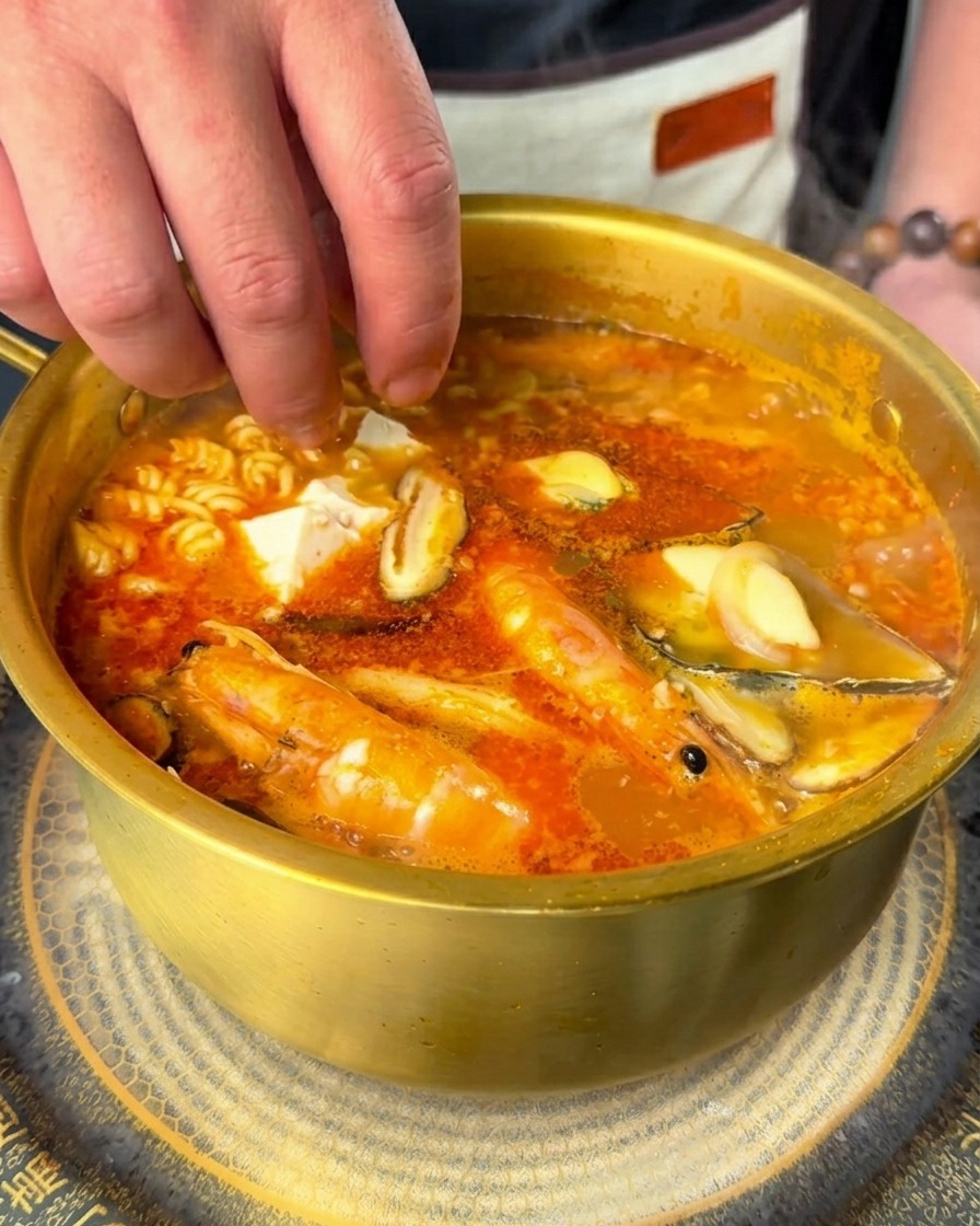 Mano che fa cadere cubetti bianchi di tofu morbido e fettine di peperoncino fresco in una pentola di ramen di mare piccante.