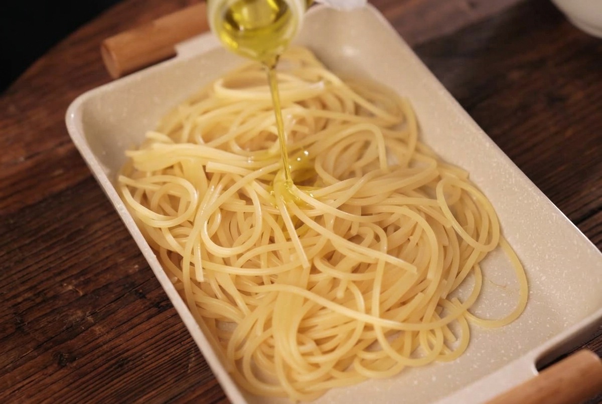 Spaghetti cotti in un vassoio color crema conditi con olio doliva dorato versato da una bottiglia.