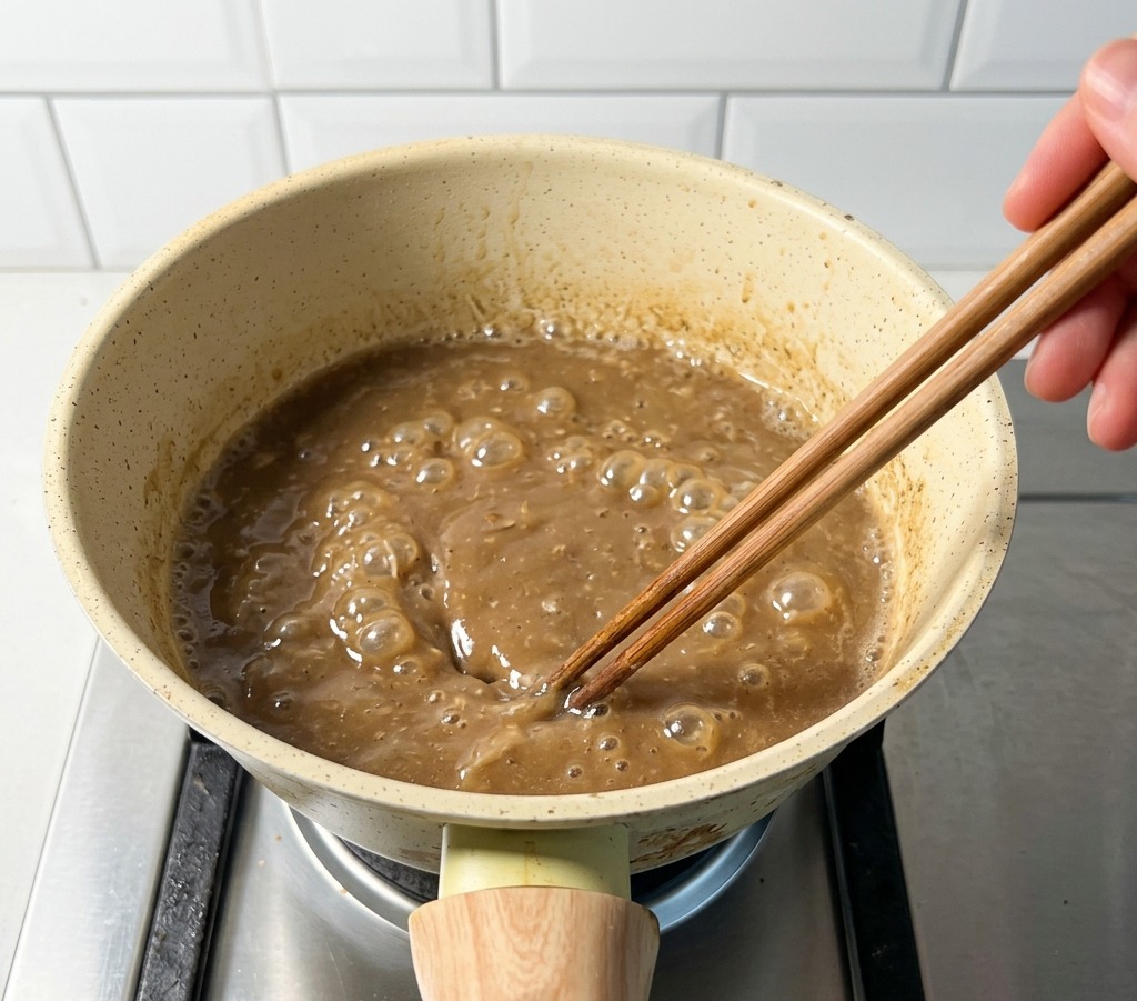 Pentolino beige su fornello a gas con brodo marrone bollente mescolato con bacchette.