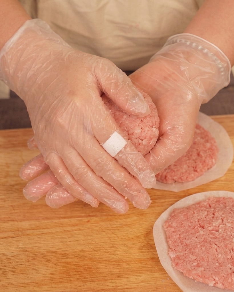Una persona con guanti trasparenti che lancia una pallina di carne macinata tra le mani sopra un tagliere.