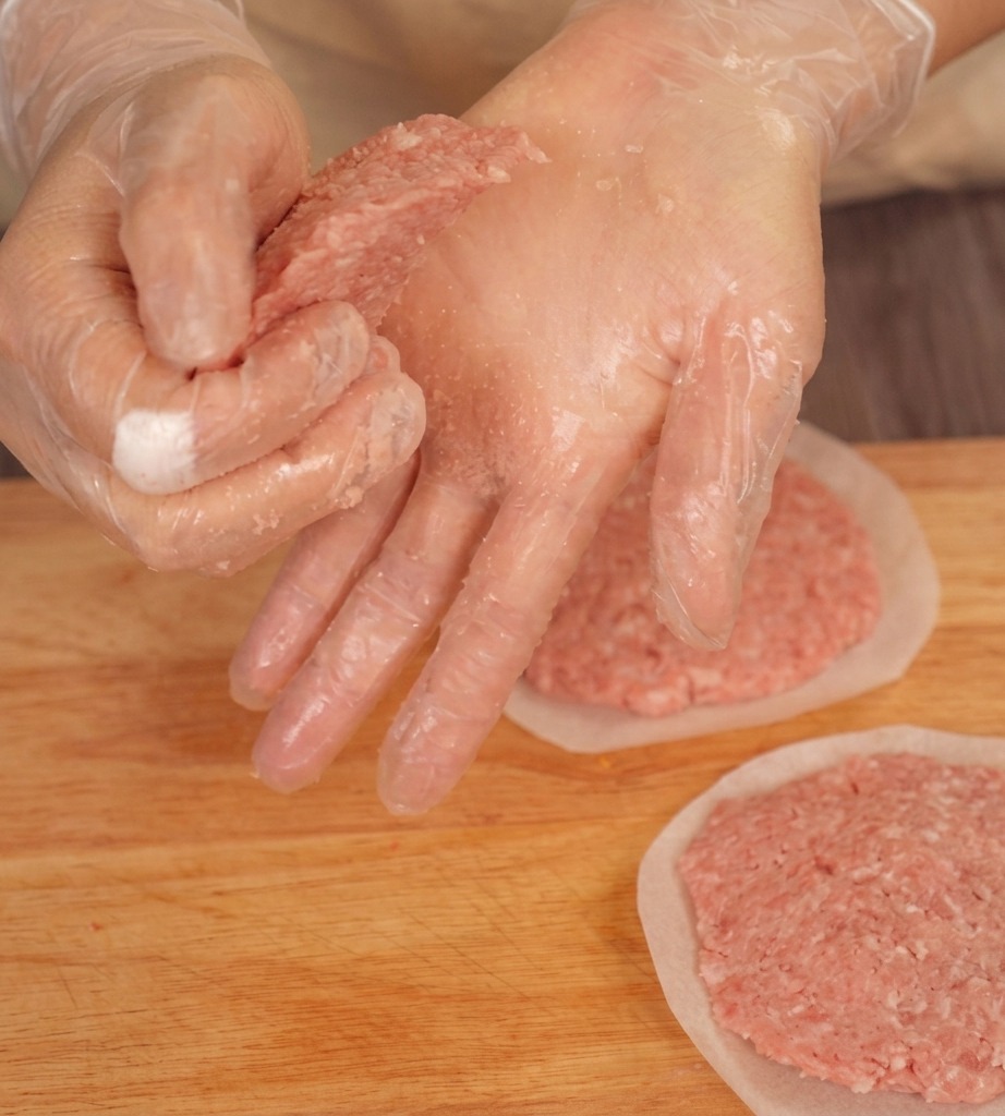 Mani guantate che premono i bordi di un patty di manzo su un tagliere per assottigliarli.