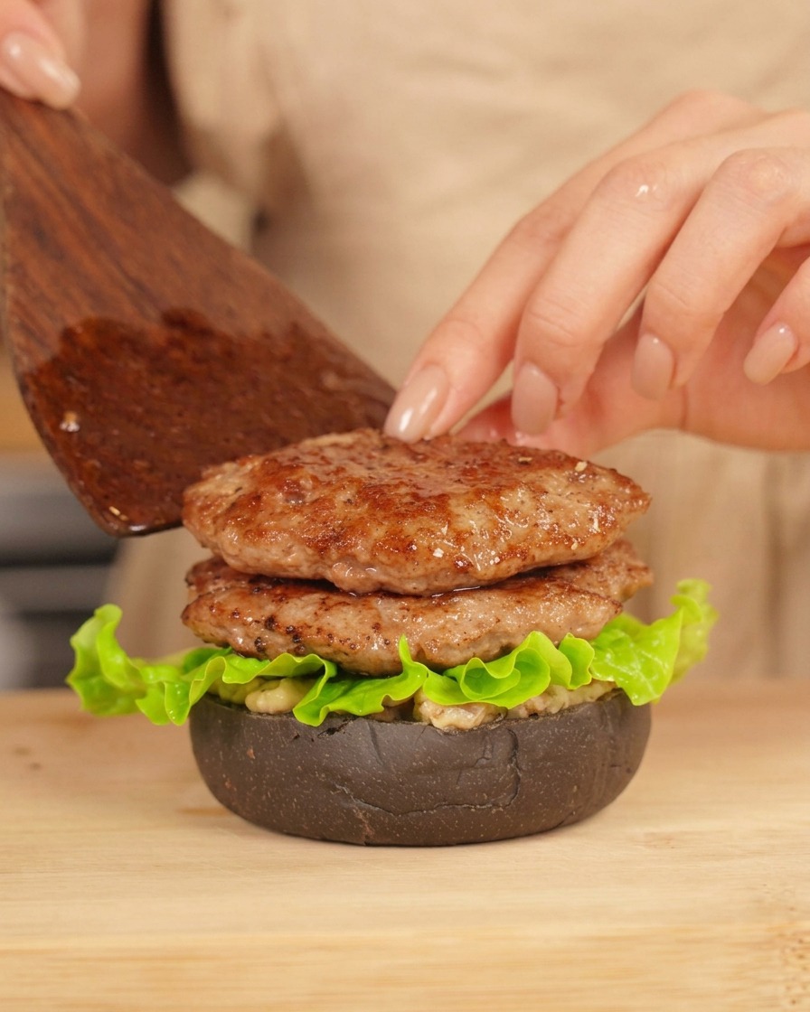 Una mano che usa una spatola per posizionare un secondo patty su un burger già assemblato con pane nero e lattuga.