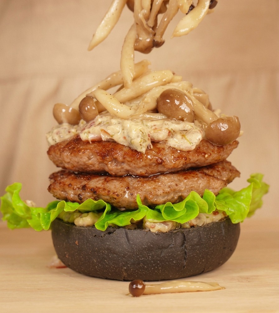 Pinze da cucina che adagiano funghi chiodini sopra i patty coperti di salsa.