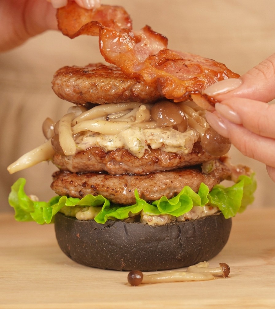 Mani che posizionano una fetta di bacon croccante sopra un alto burger doppio assemblato.