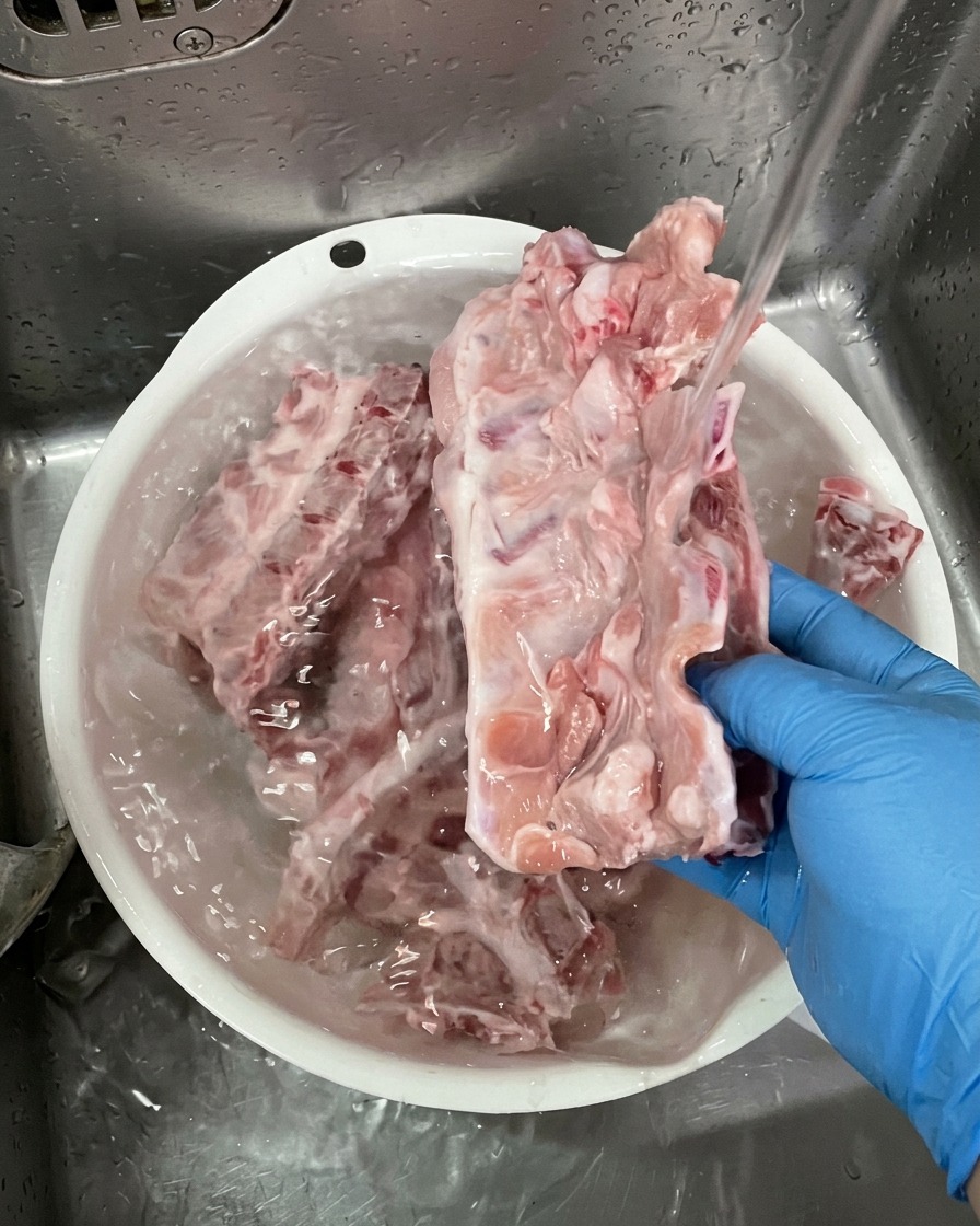 Una persona con guanti blu lava grandi ossa di maiale crude sotto lacqua corrente in una bacinella di plastica bianca.
