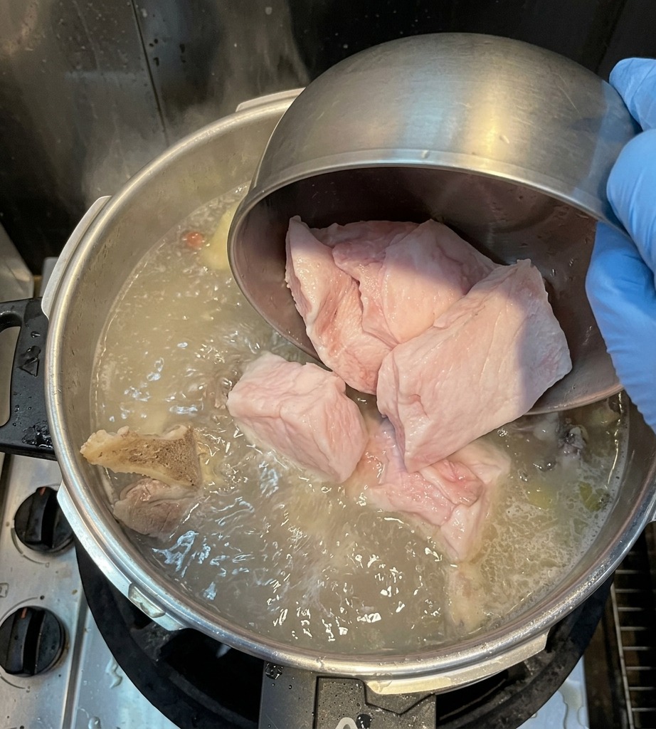 Una mano con un guanto versa grandi pezzi di lardo di maiale bianco da una ciotola di metallo in una pentola di brodo in ebollizione.