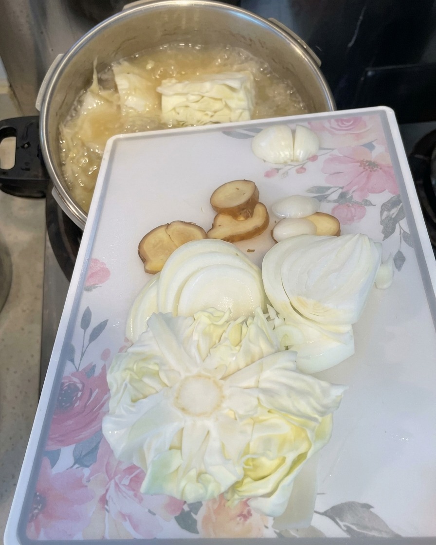 Un tagliere bianco con cipolle affettate, zenzero e cavolo viene inclinato per far scivolare gli ingredienti nella pentola in ebollizione.