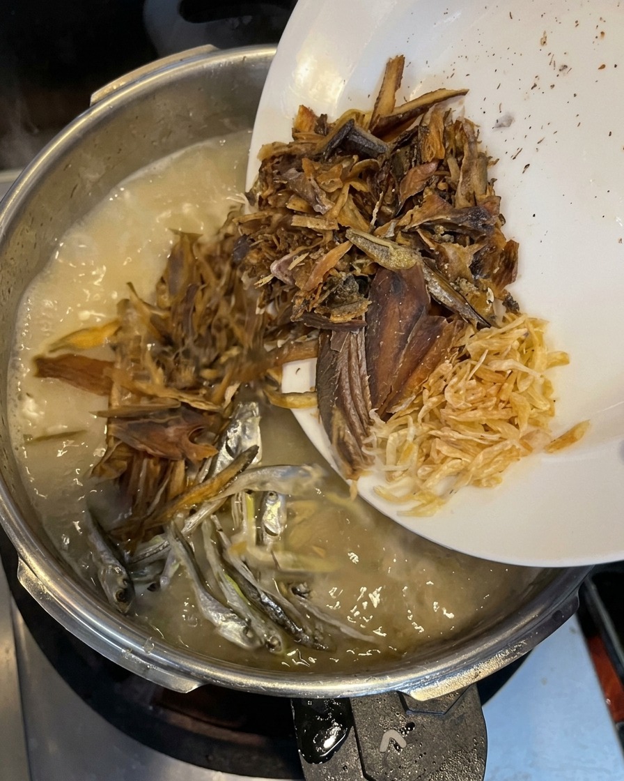 Fiocchi di pesce essiccato, piccole sardine argentate e minuscoli gamberetti essiccati vengono versati da un piatto bianco nel brodo cremoso in ebollizione.