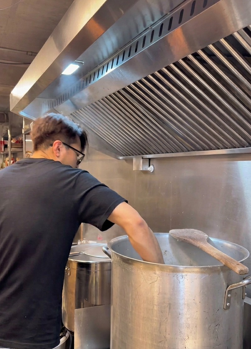 Uno chef in maglietta nera si china su una grande pentola di metallo filtrando un brodo bianco e cremoso attraverso un colino.