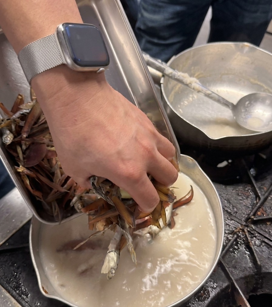 La mano di uno chef aggiunge una manciata di ingredienti essiccati di colore marrone scuro in una pentola con del brodo cremoso.