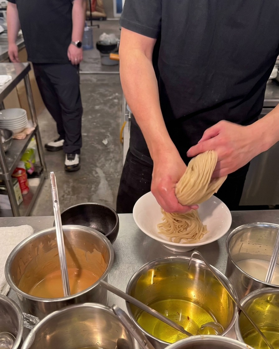 Uno chef usa entrambe le mani per piegare e sistemare un nido di noodles spessi e chiari in una ciotola di ceramica bianca.