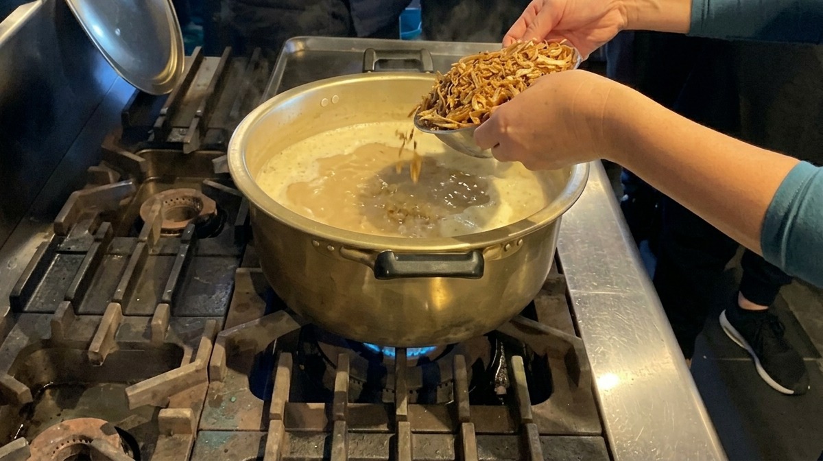Mani che aggiungono una manciata di acciughe essiccate in una grande pentola di ottone piena di zuppa bianca e cremosa che bolle.