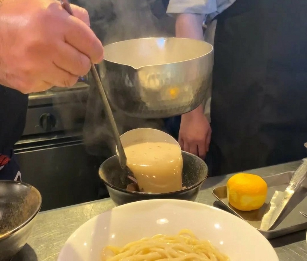Brodo per ramen di pesce denso e cremoso versato da un mestolo di metallo in una ciotola di ceramica nera.