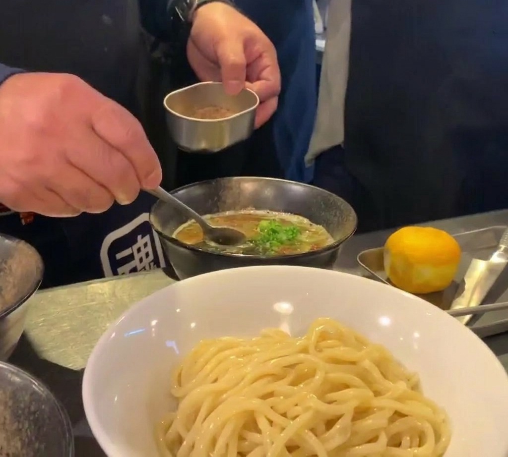 Un cuoco che usa un piccolo contenitore metallico e un cucchiaio per spolverare della fine polvere di pesce marrone su una ciotola di zuppa scura e densa.