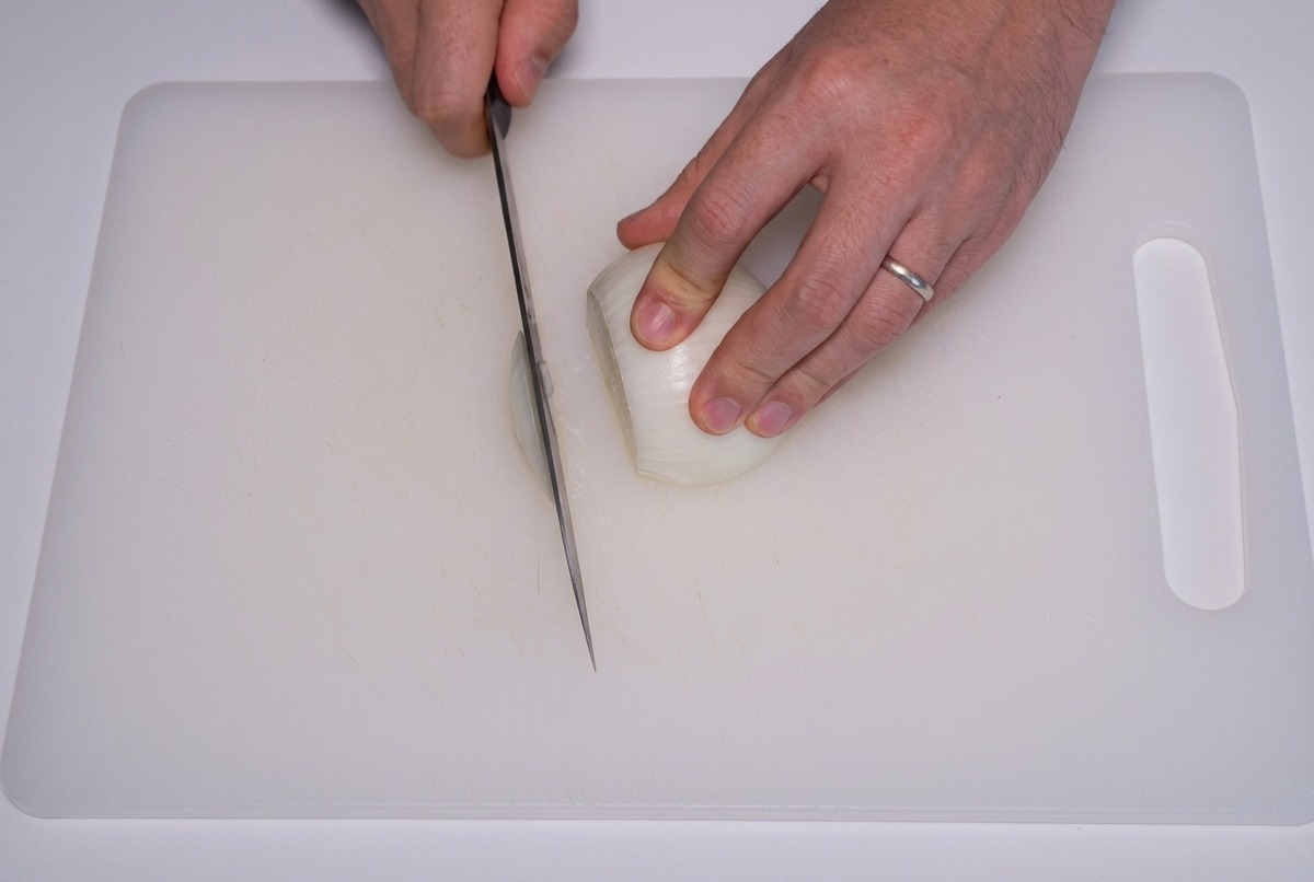 Mani che usano un coltello affilato per tagliare una cipolla bianca a strisce sottili su un tagliere bianco.