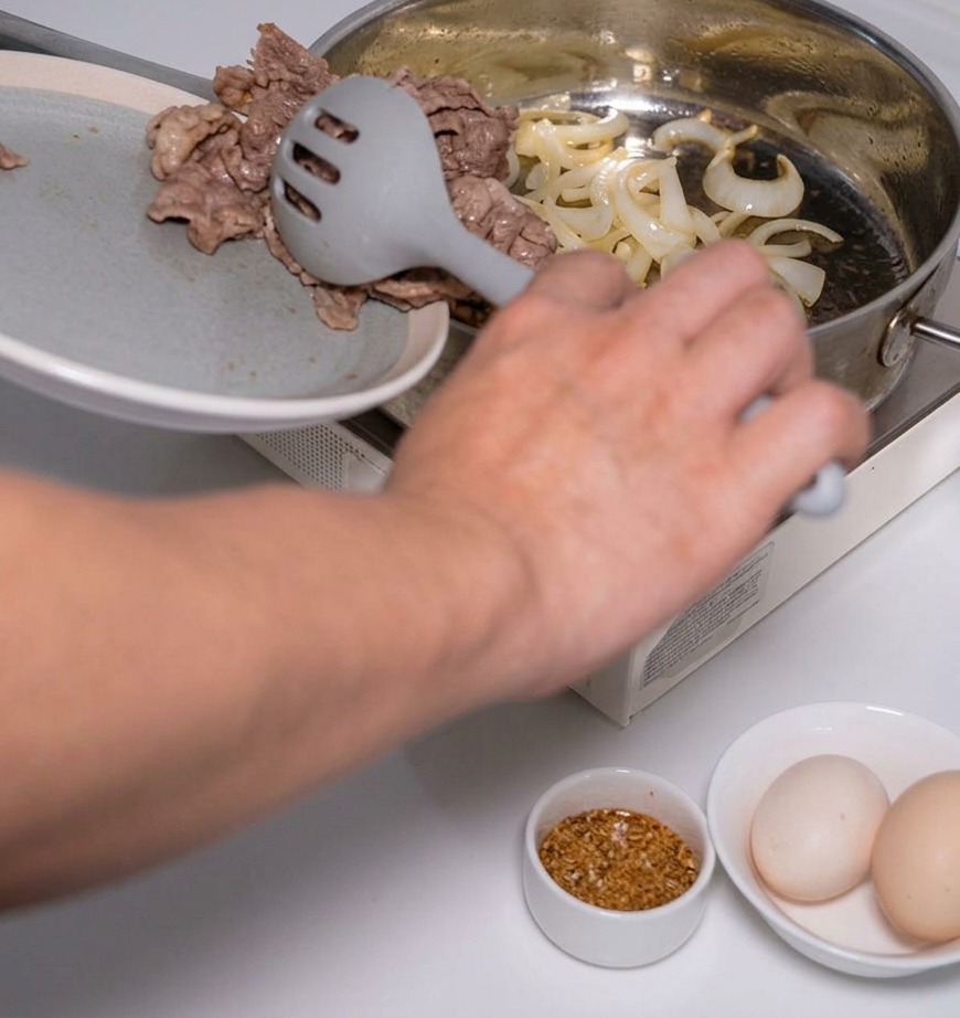 Una persona che usa una schiumarola grigia per aggiungere fette di manzo cotte nella padella con le cipolle soffritte.