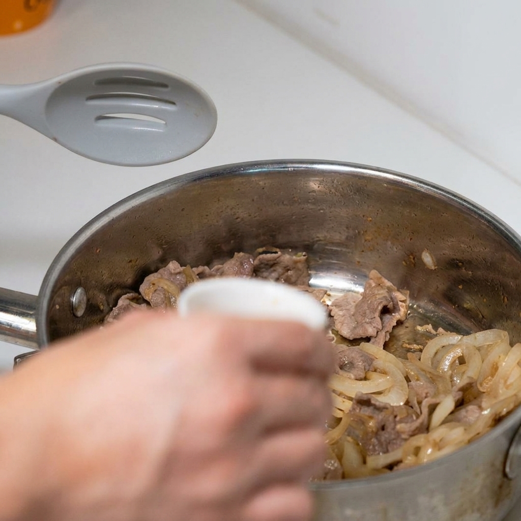 Una mano che versa una ciotolina di salsa preconfezionata sul manzo e sulle cipolle in padella.