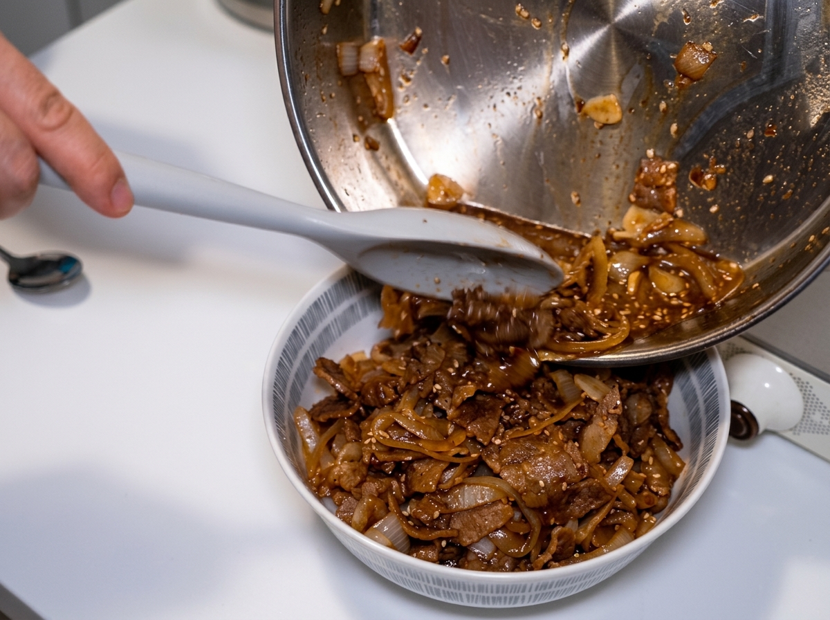 Una padella inclinata e una spatola che versano le cipolle rimaste e il ricco brodo scuro su una ciotola di riso e manzo.
