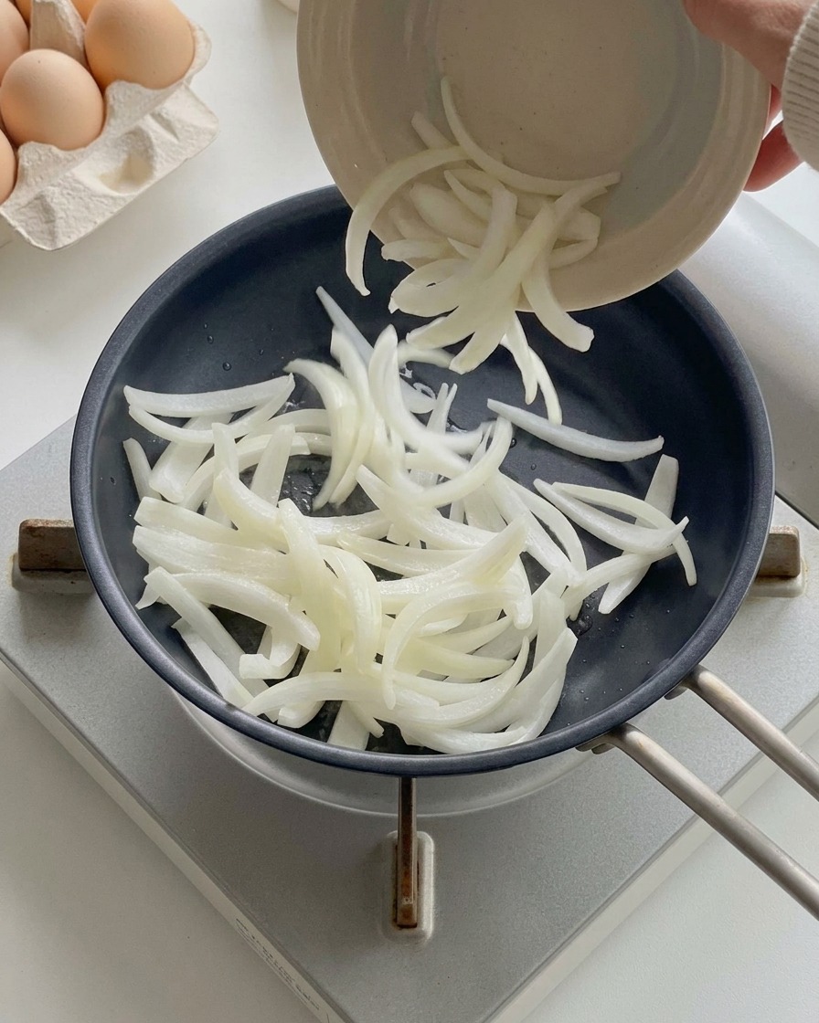 Cipolle bianche affettate versate da una ciotolina in una padella nera già calda.