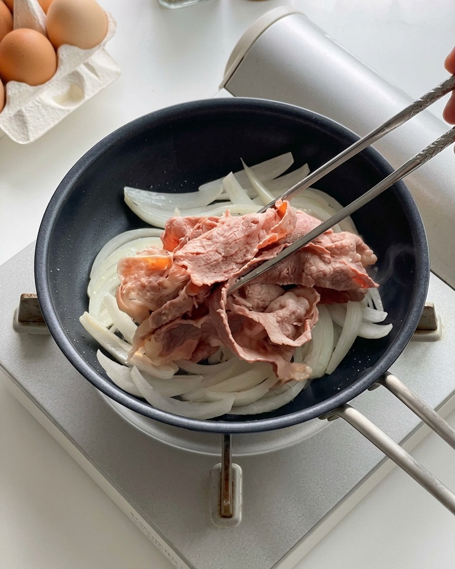 Bacchette di metallo che adagiano le fette di manzo sbollentate sopra le cipolle in cottura in una padella nera.