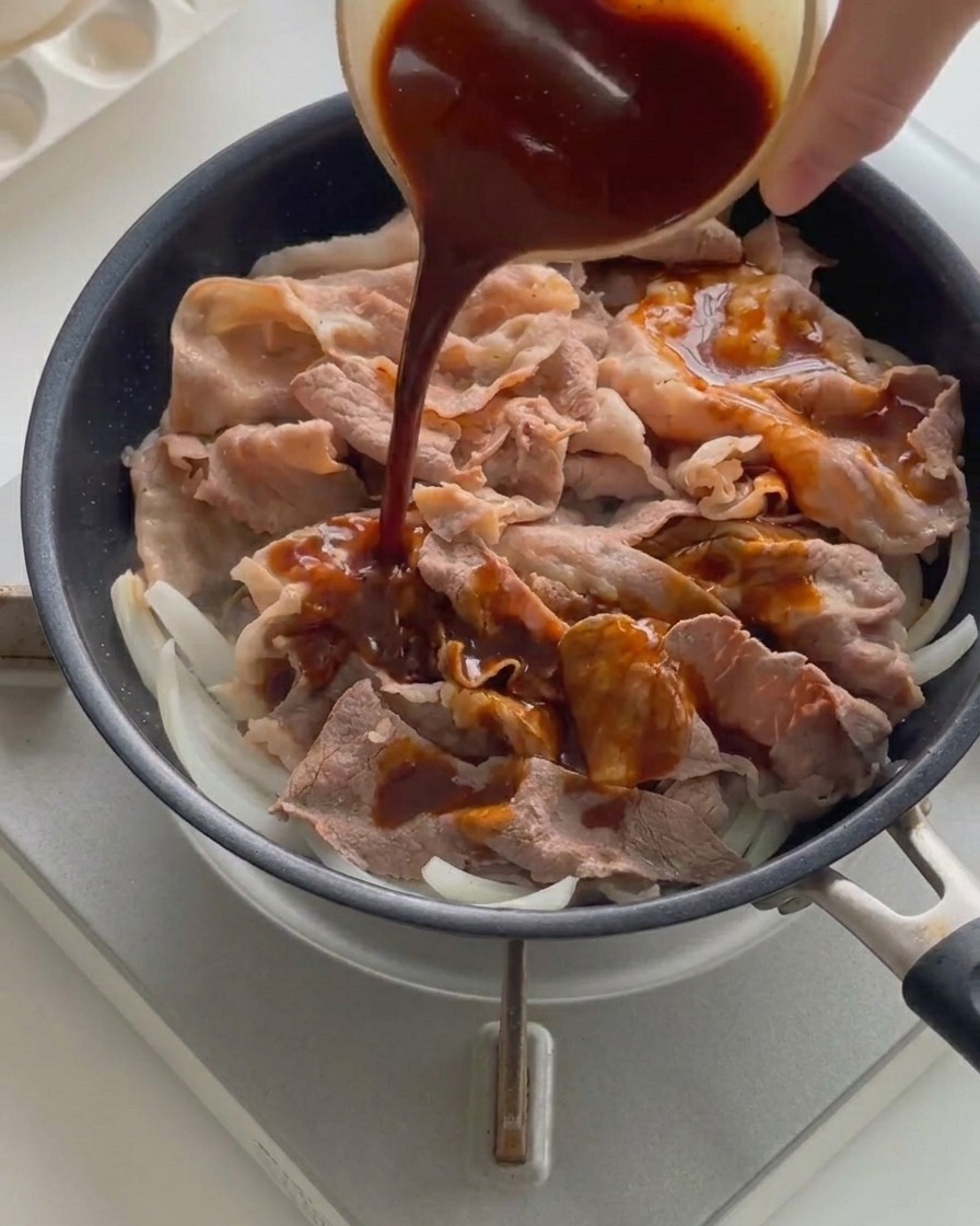 Una salsa liquida scura versata da una ciotolina sopra le fette di manzo cotto e le cipolle in una padella calda.