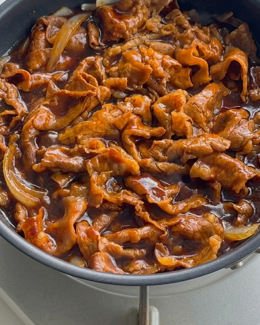 Primo piano di straccetti di manzo e cipolle che sobbollono in una salsa densa, scura e lucida in una padella.