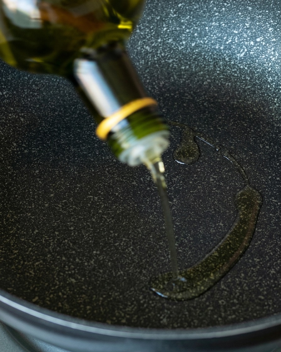 Olio di avocado limpido versato da una bottiglia di vetro verde in una padella antiaderente scura e picchiettata.