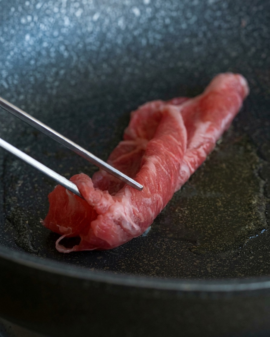 Pinze di metallo che adagiano una fetta sottile e marezzata di manzo crudo in una padella calda con olio luccicante.