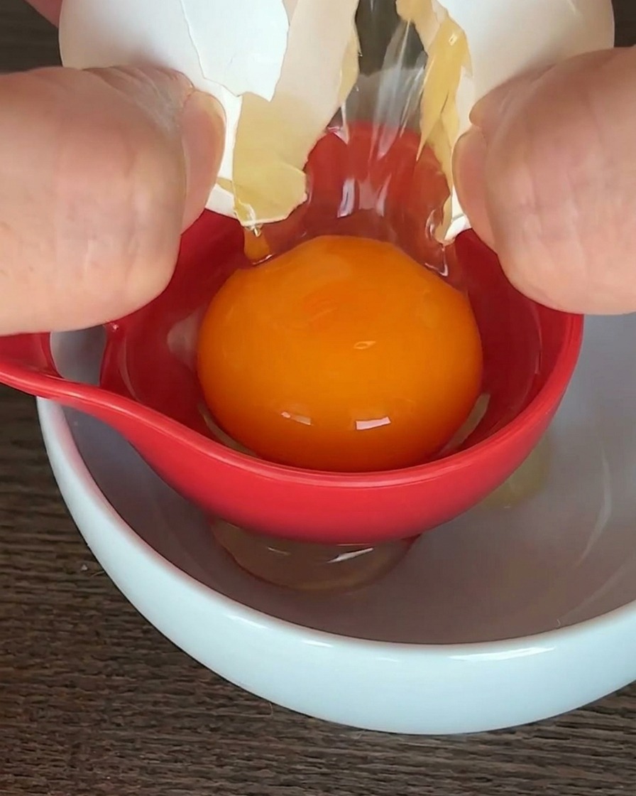 Un uovo crudo separato con un utensile rosso sopra una ciotola bianca, con il tuorlo arancione al centro.