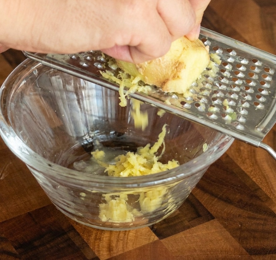 Una mano grattugia un pezzo di zenzero fresco su una grattugia di metallo, facendo cadere la pasta in una ciotolina di vetro.
